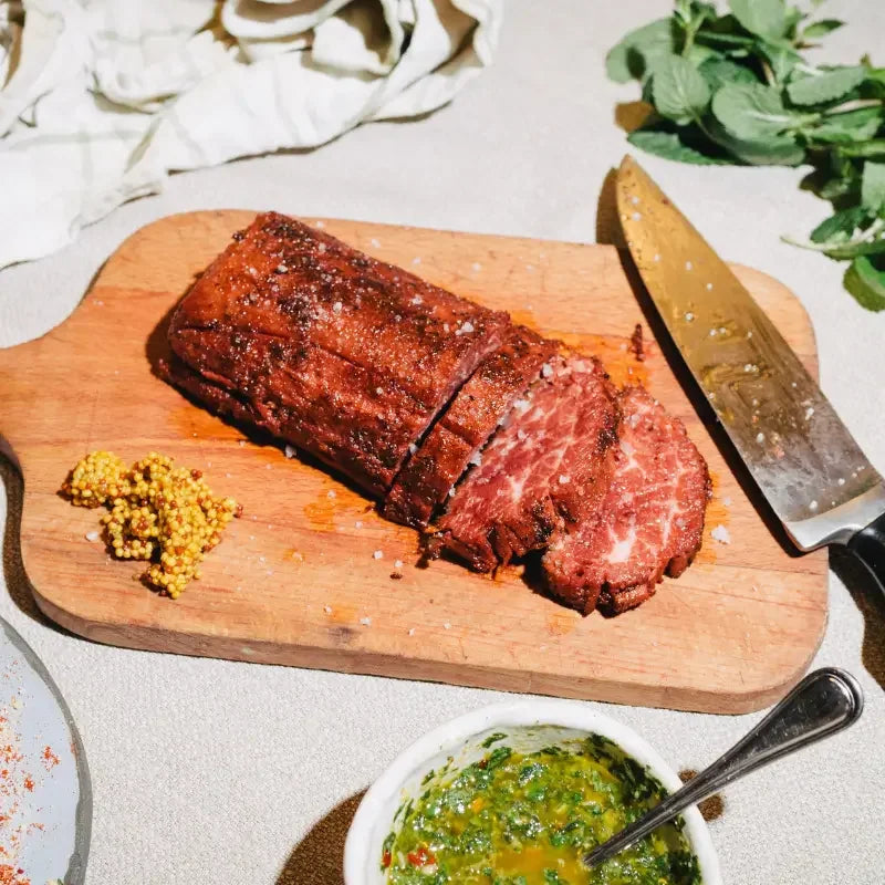 whole cut loin steak on a wooden cutting board with a knife, herbs, and a bowl of green sauce.